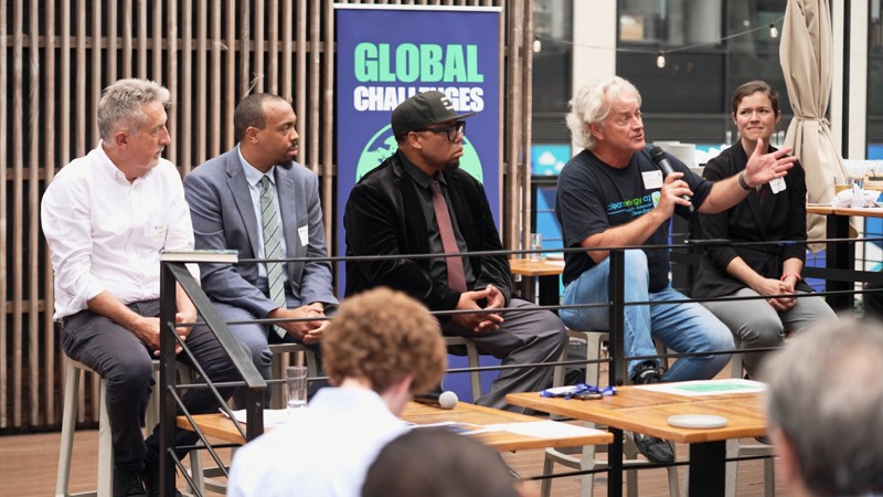 Five panelists seated in front of a banner, one holding a microphone